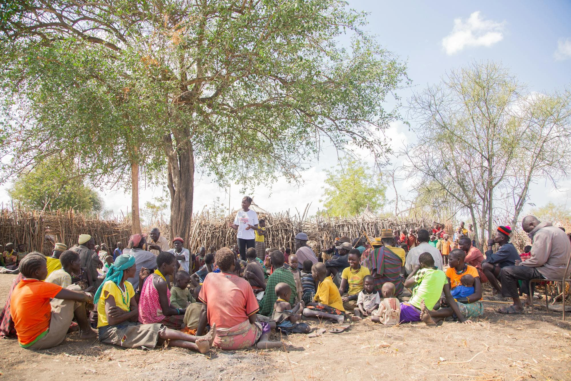 A diverse group of adults and children meeting outdoors under a tree