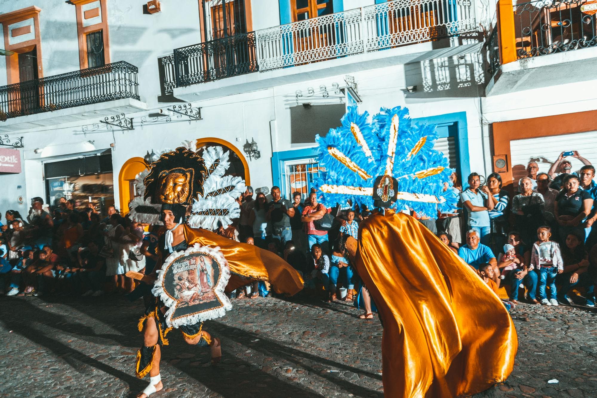 A traditional dancer in feathered headdress and gold cape performing in a Mexican town square at night.