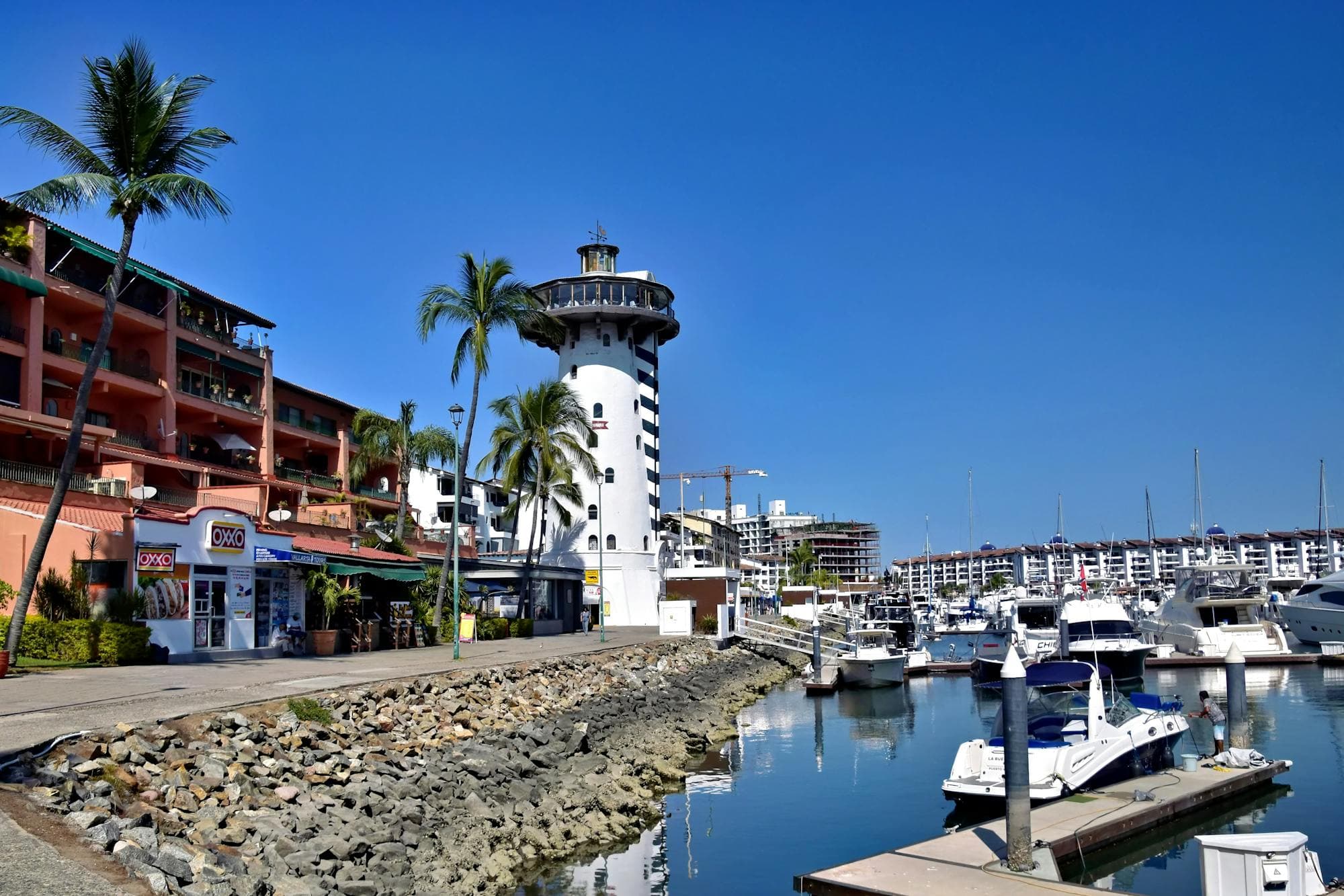 The Marina Vallarta lighthouse and harbor pier under a midday sky, with the marina stretching to the mountains beyond.
