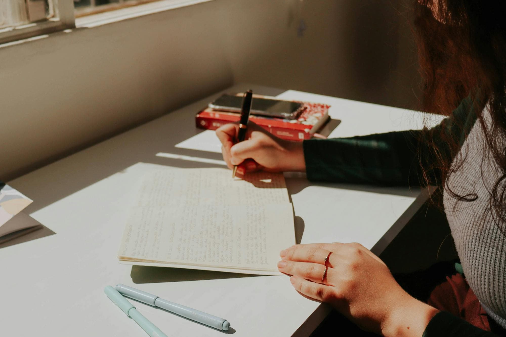 A hand writing in a notebook by a sunlit window in warm afternoon light.